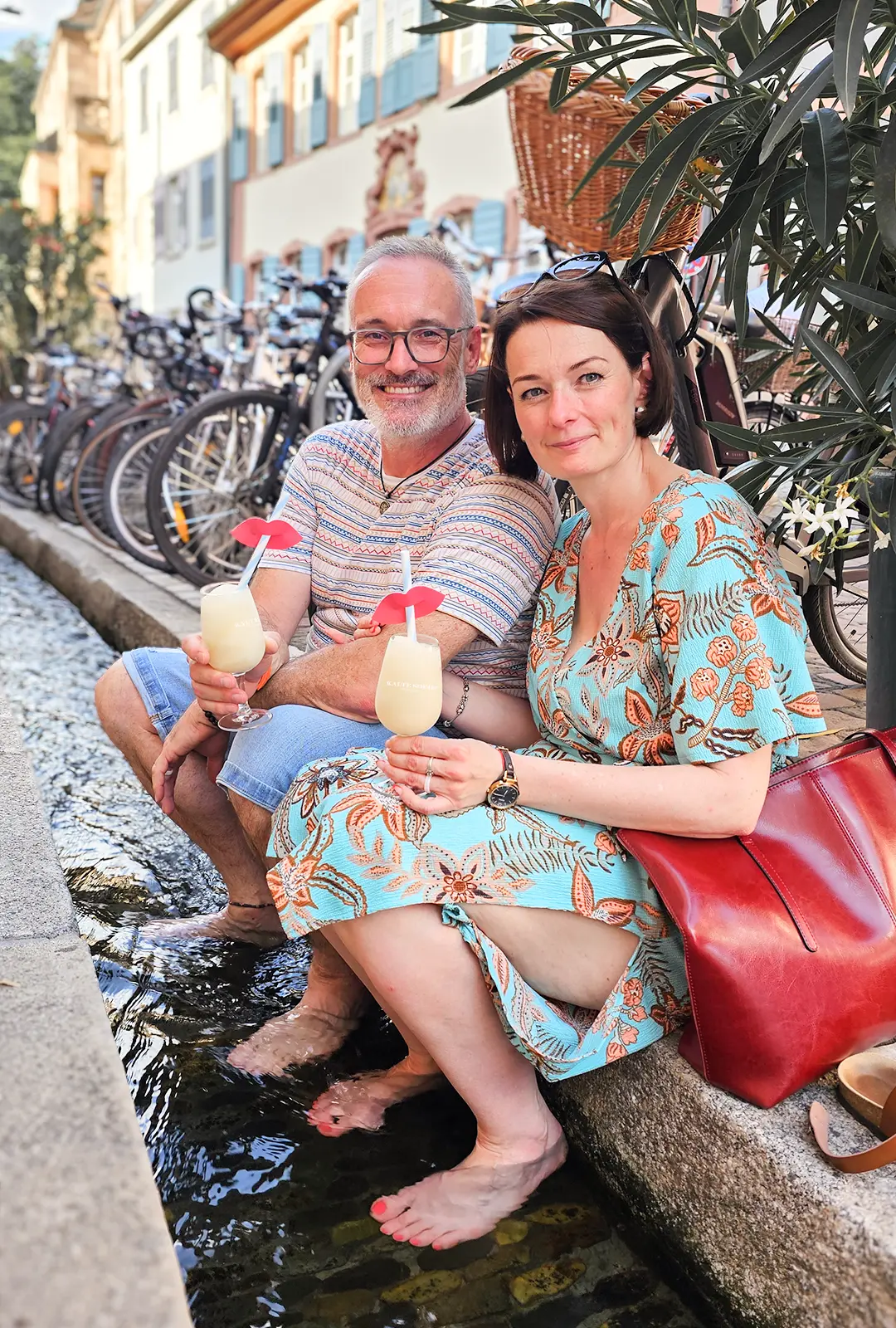 Ein Mann und eine Frau sitzen lächelnd nebeneinander in der Freiburger Altstadt und kühlen ihre Füße im Bächle. Beide halten die Klate Sofie Freiburg in den Händen.