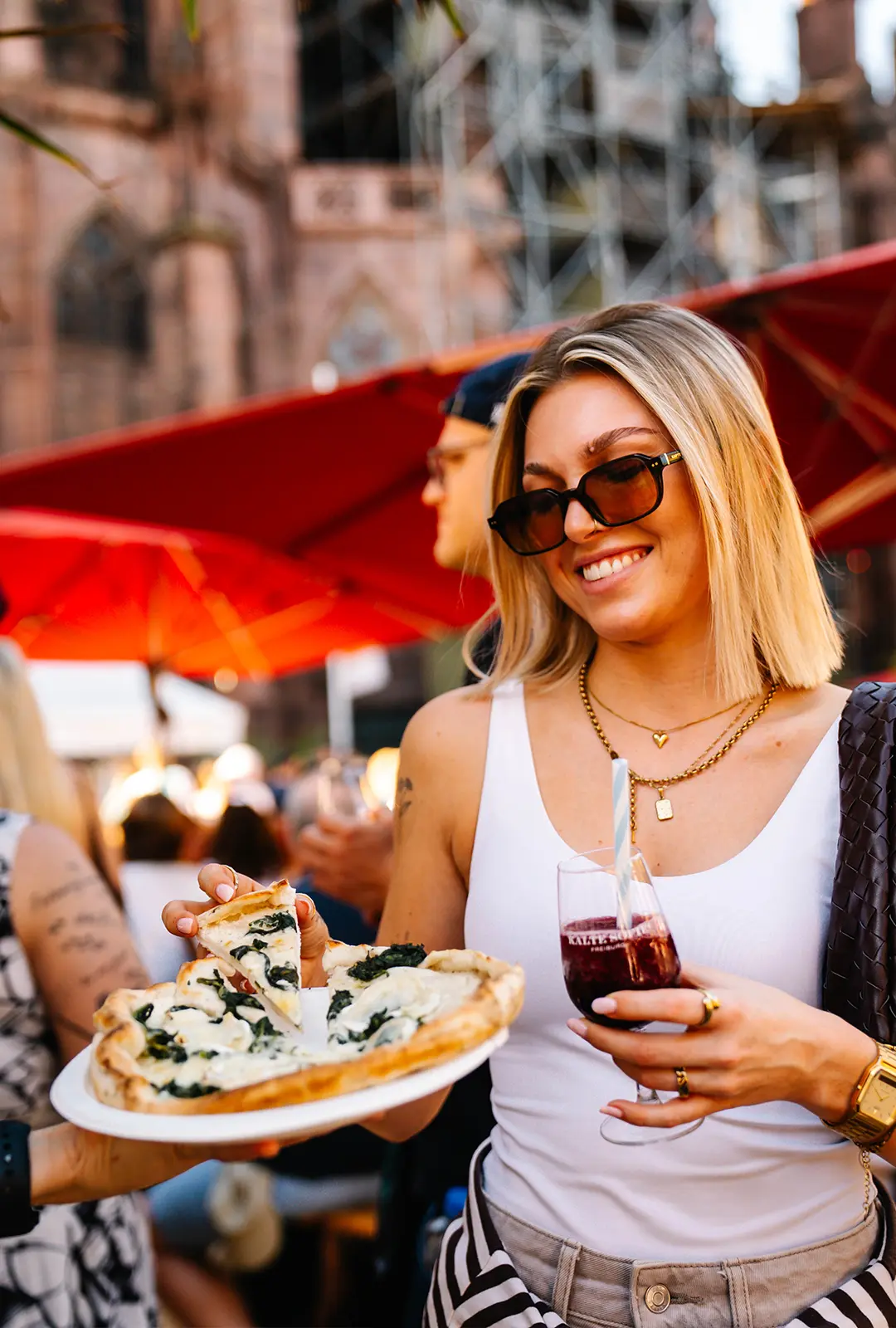 Eine Frau hält ein Glas „Kalte Sofie Freiburg" in der Hand und nimmt sich mit der anderen Hand ein Stück Pizza.
