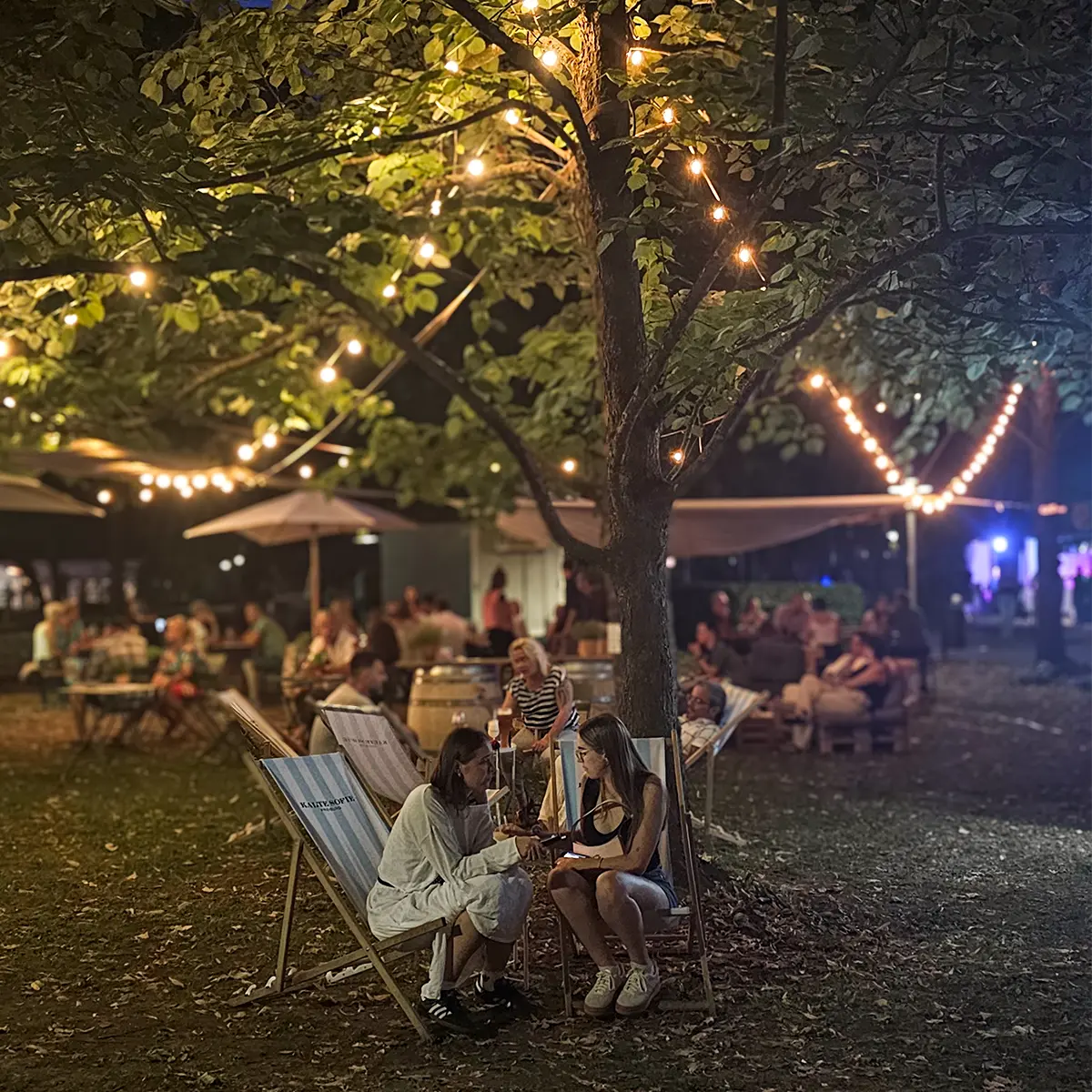 Zwei Frauen sitzen in blau-weiß gestreiften Liegestühlen der „Kalten Sofie" bei einem Fest in stimmungsvoller Abendatmosphäre. Die umliegenden Bäume sind mit Lichterketten geschmückt.
